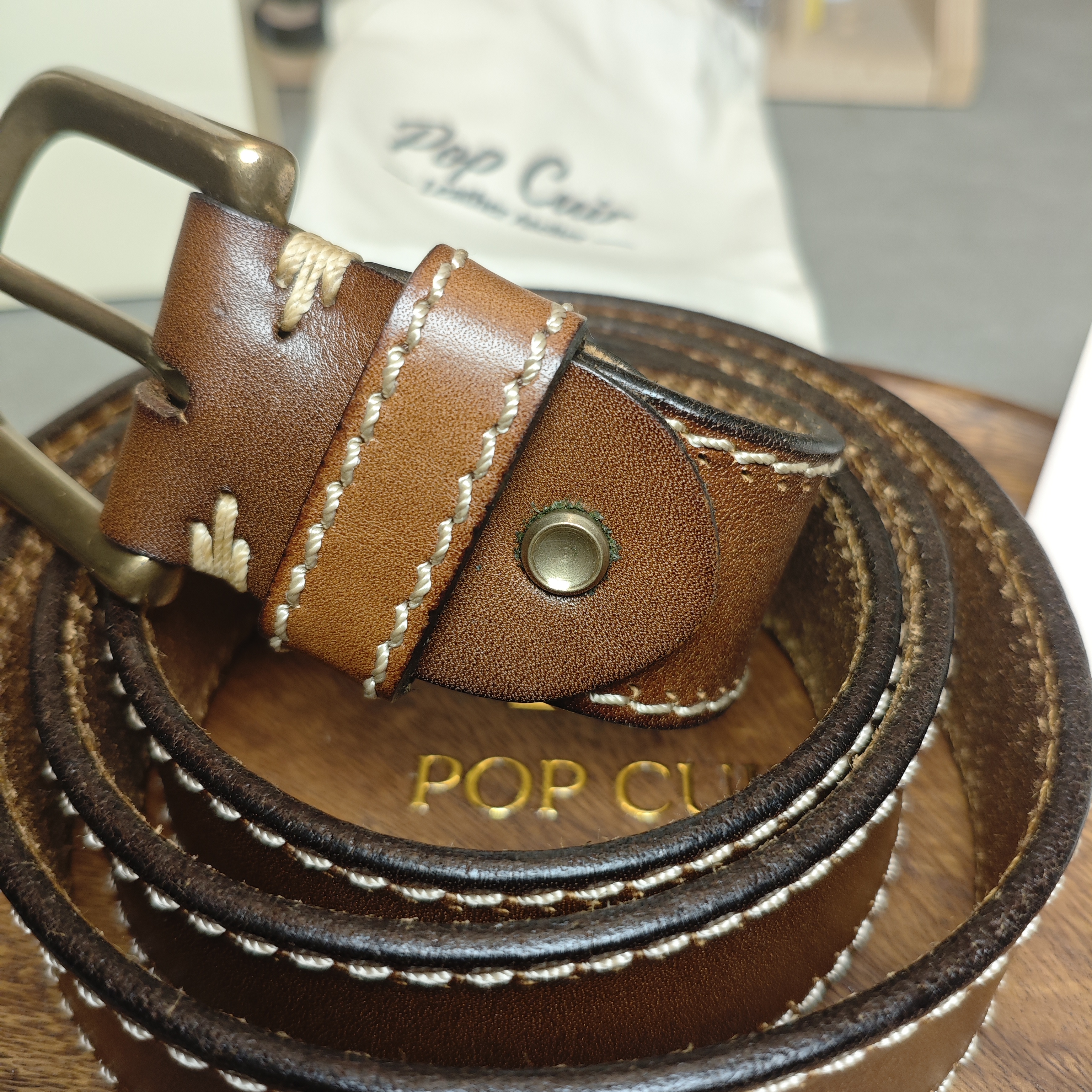 Close-up detail of Tan color leather belt showing hand-aged brass pin buckle and white contrast double-stitch on rustic wood background