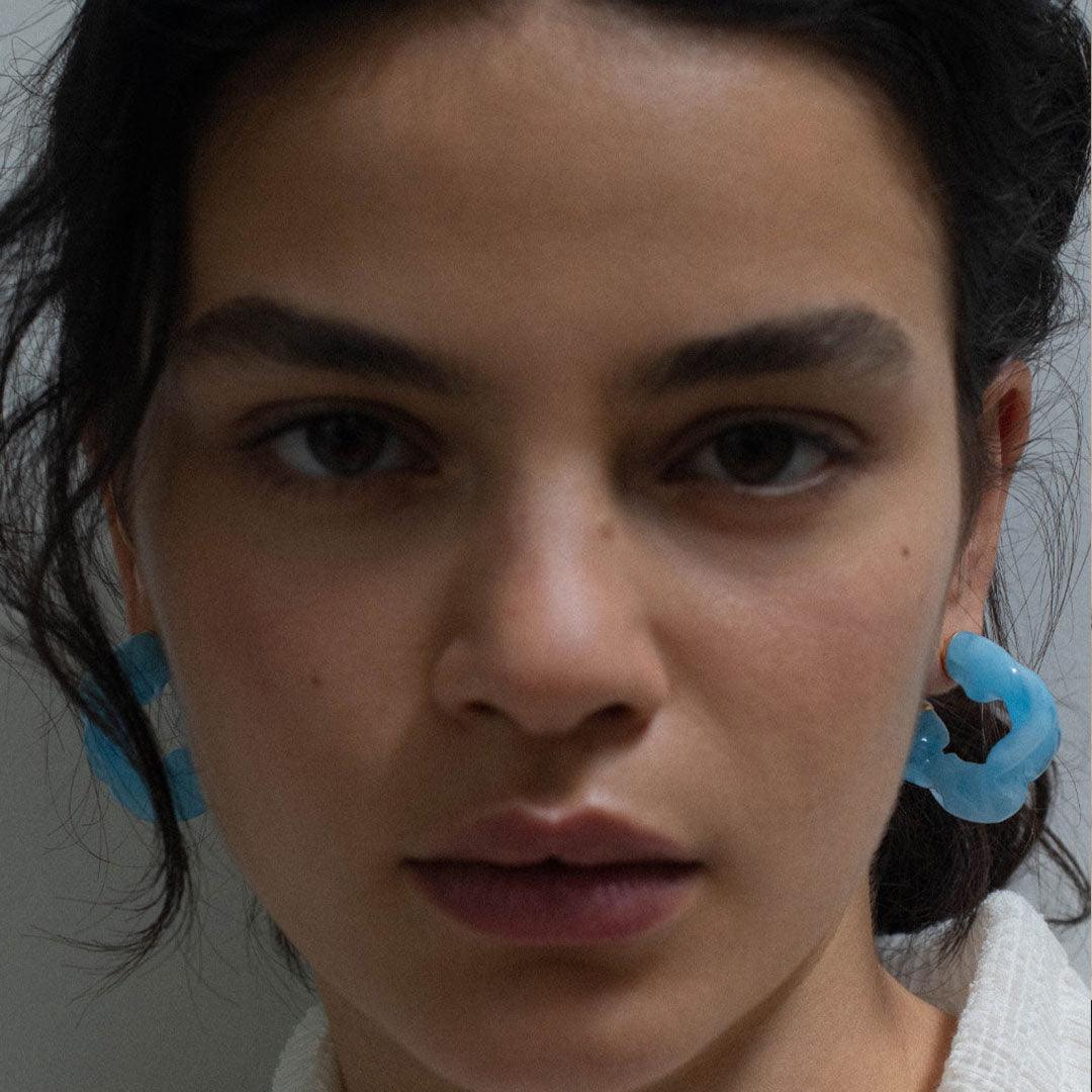 CULT STATEMENTPIECES BLUE BIO RESIN GOLD VERMEIL EARRINGS featuring a clean chic hoop design and modern jelly blue color ,perfect for dailylife/party/office/casual occasion. Displayed in a cool  lifestyle setting with a model looking straight at the people.