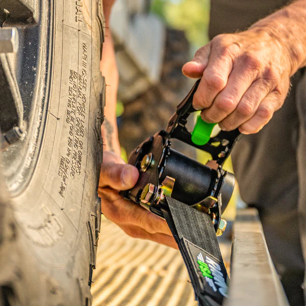 A person&rsquo;s hands tighten a ratchet strap around a vehicle tire, securing it to a trailer. The focus is on the strap mechanism and the tire tread.