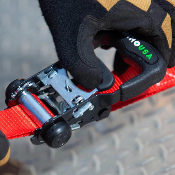 A gloved hand tightens a red ratchet strap using a metal and black plastic ratchet handle marked USA on a textured metal surface.