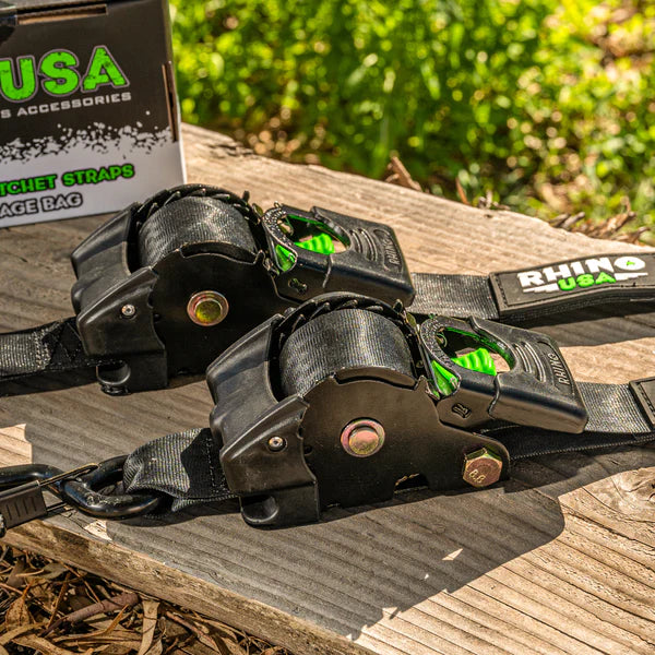 Two black Rhino USA ratchet straps with green accents are placed on a wooden surface outdoors. A product box is partially visible in the background.