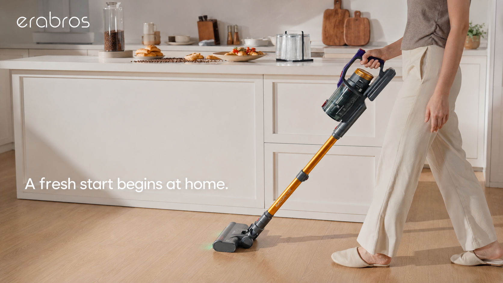 erabros lifestyle scene: A woman uses a gold and black cordless stick vacuum to clean a bright modern kitchen floor. The white island counter features coffee beans and pastries, with the text "A fresh start begins at home."