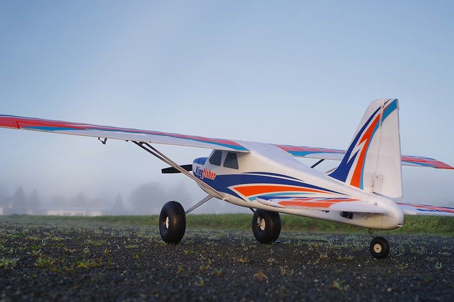 FMS 1400mm Kingfisher PNP model airplane with wheels and colorful racing stripes on a misty field.