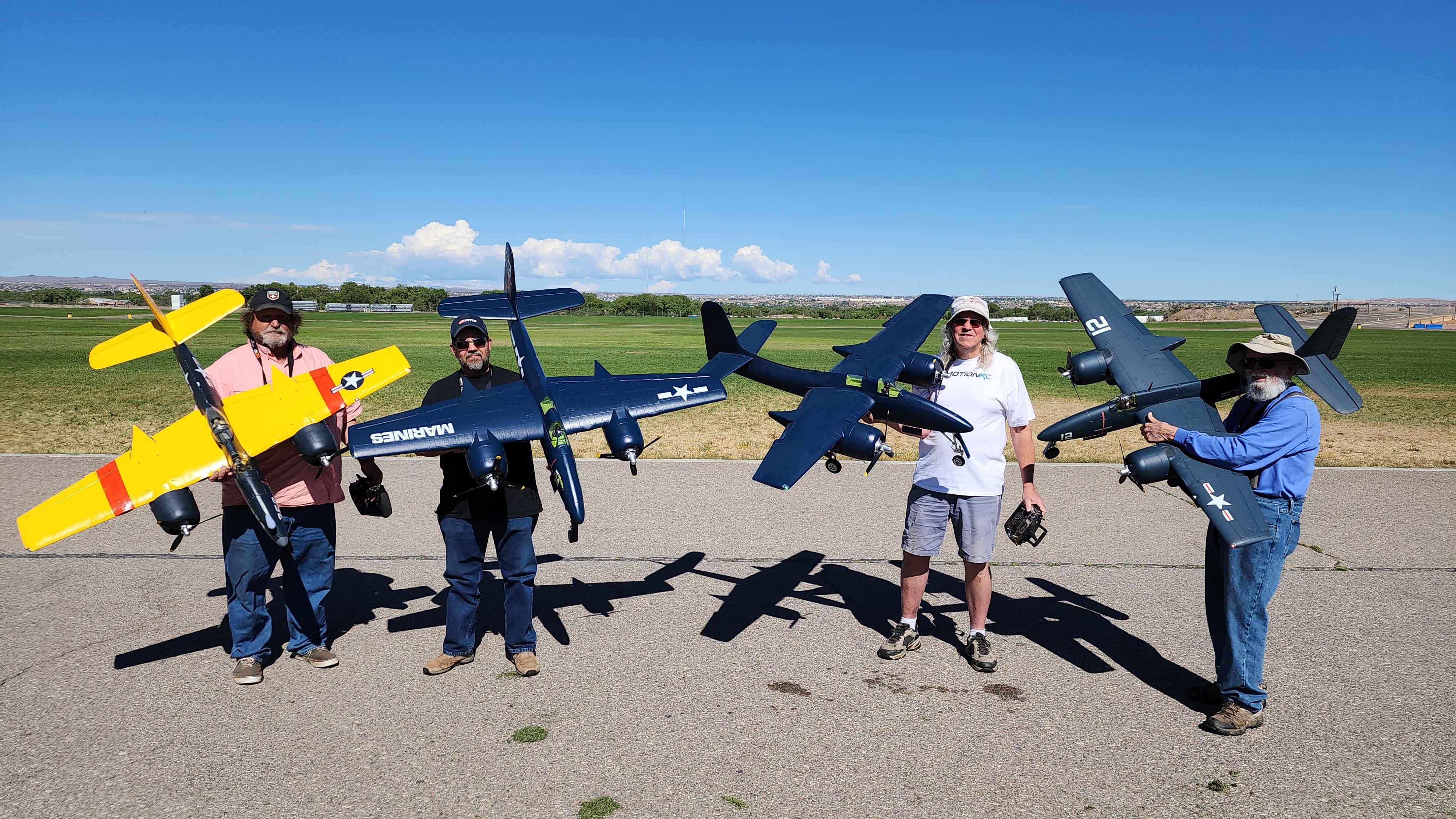 Four individuals holding large RC airplanes, including a bright yellow model and several dark blue models with "MARINES" markings, standing on a paved runway with a grassy field and clear blue sky in the background.