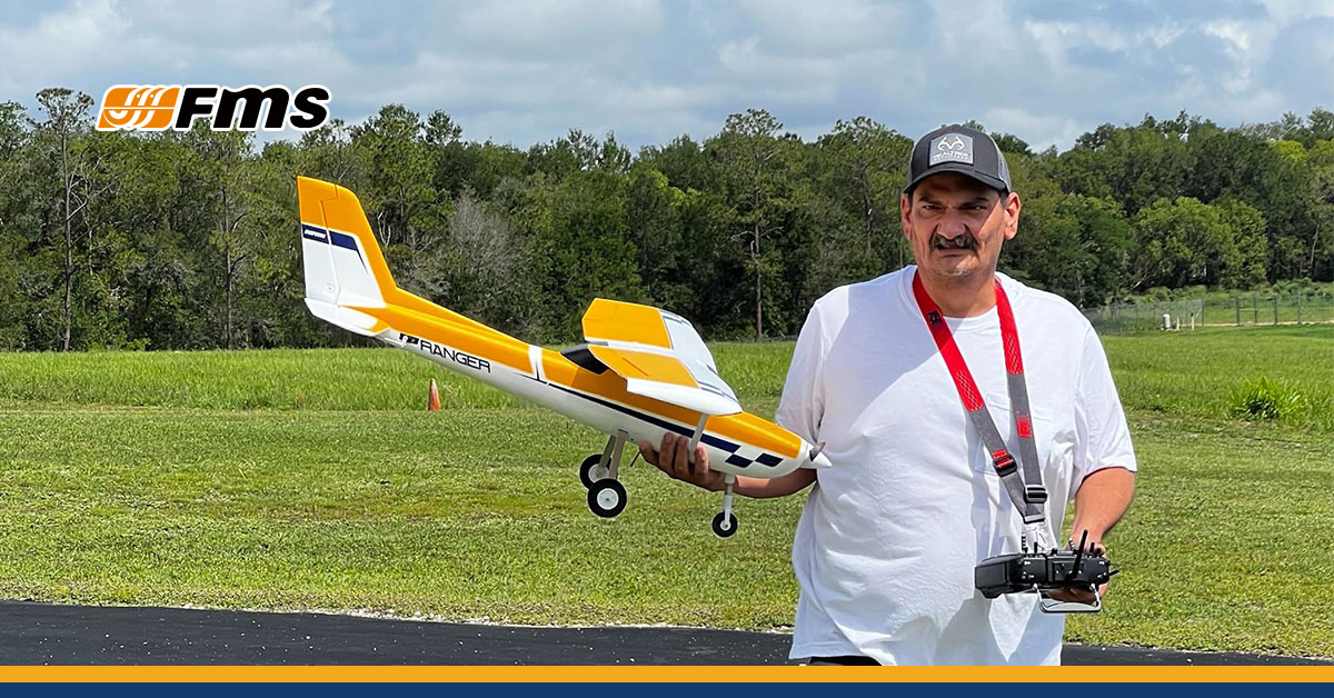 A man holding an FMS Ranger R/C airplane with a yellow and white color scheme at an outdoor flying field.