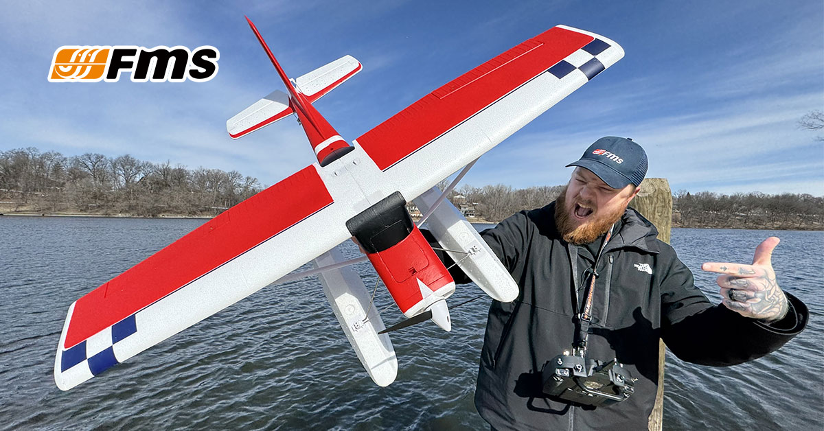 A man holding a red and white FMS R/C airplane model outdoors near a lake.