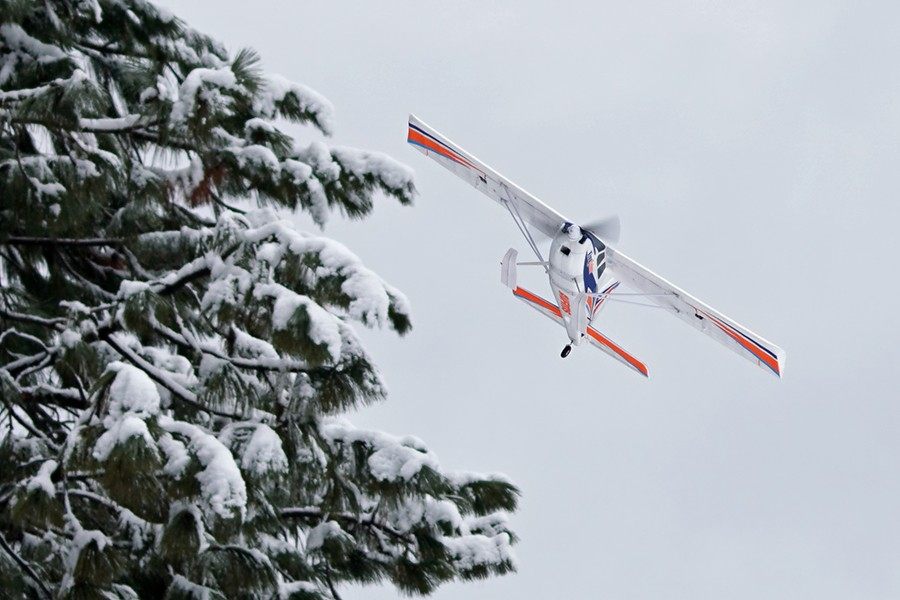 FMS 1400mm Kingfisher PNP aircraft in flight, banking left near snow-covered pine trees.