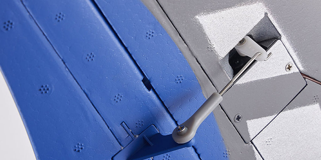 Close-up of a control linkage connected to a servo arm on the blue and gray wing of a remote control model airplane.