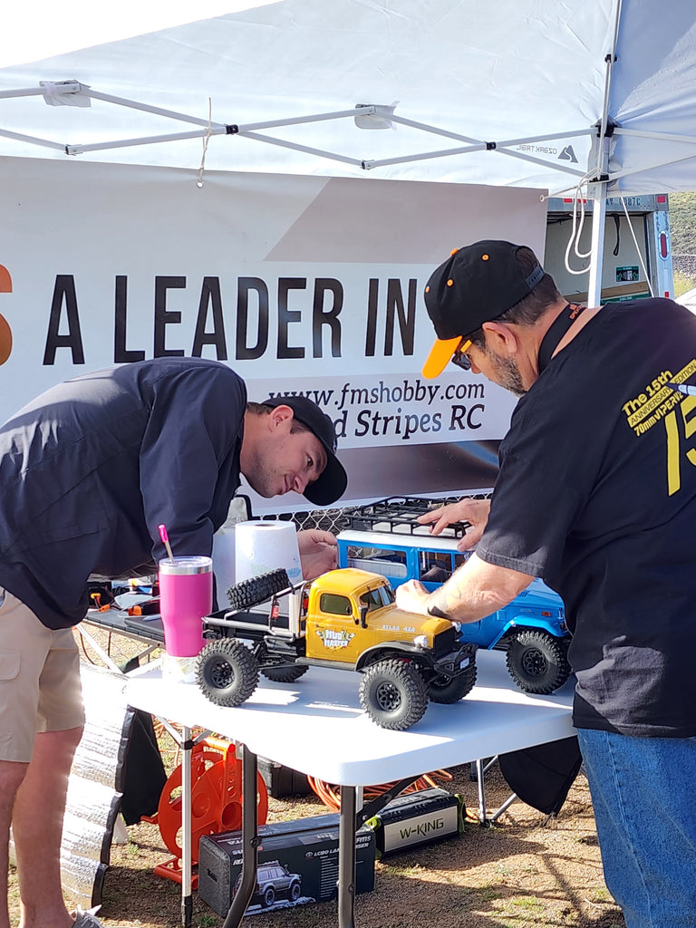 Two enthusiasts at an event meticulously adjust their Fmshobby RC Trucks, including a yellow mud master model