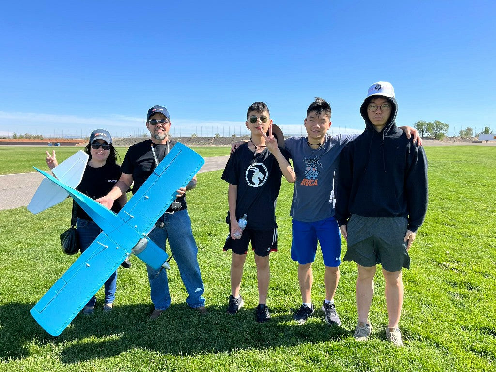 A group of individuals standing on a grassy field holding a large blue RC airplane, with a clear blue sky and a runway in the background.
