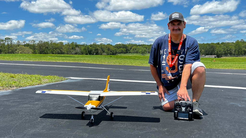 Person kneeling on asphalt runway holding a remote control next to a yellow and white remote control model airplane with green grass and blue sky in the background.