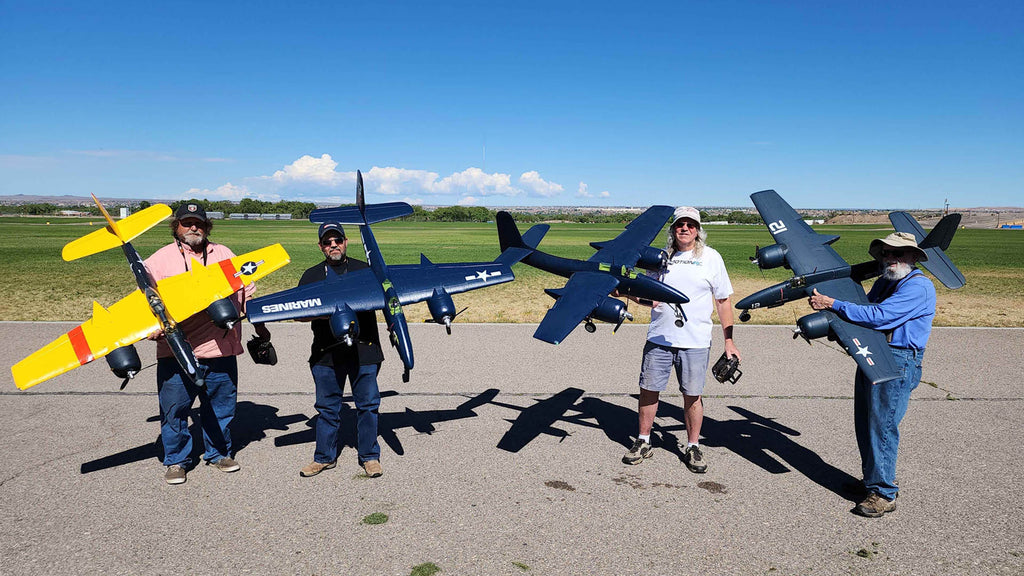 Four people standing on a runway holding large remote control model airplanes painted in yellow and dark blue with U.S. Navy and Marines insignia, with green grass and blue sky in the background.