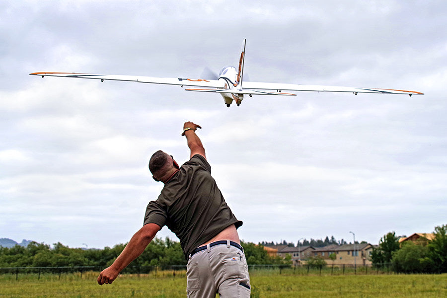 FMS 3000mm Fox aerobatic glider being thrown by a man in a grassy field under a cloudy sky. White and orange glider soaring.