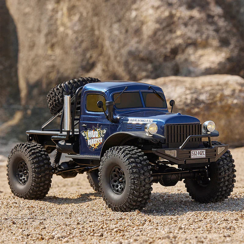 Blue vintage-style RC truck with rugged tires, spare wheel, and "Mud Master" decal, parked on sandy terrain near rocks.