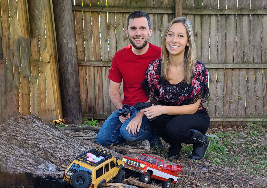A smiling couple enjoys a day outdoors, posing with their favorite Fmshobby RC Trucks and handheld controllers