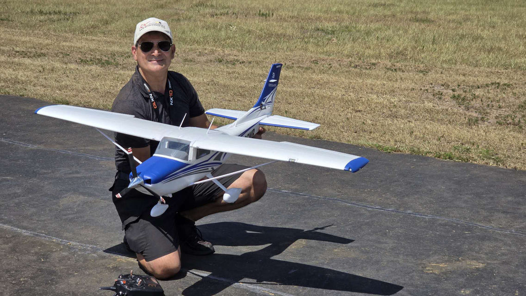 Man kneeling on a runway holding a blue and white remote control model airplane outdoors on a sunny day.