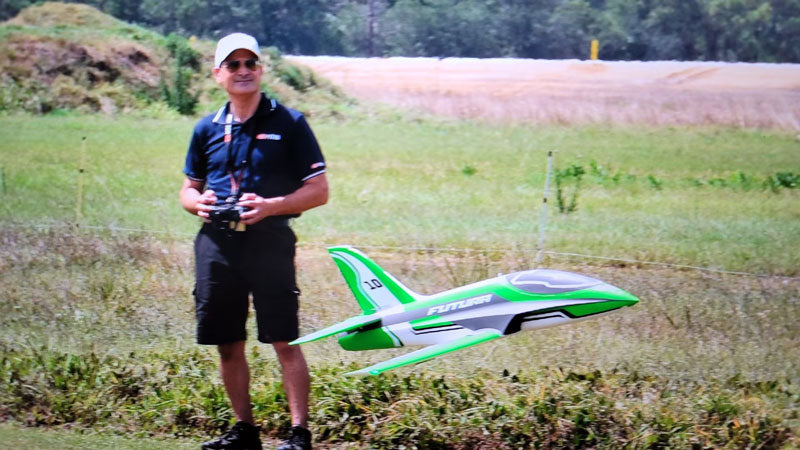 Man standing on grass holding a remote controller, flying a green and white remote control model airplane numbered "10" in an outdoor field.