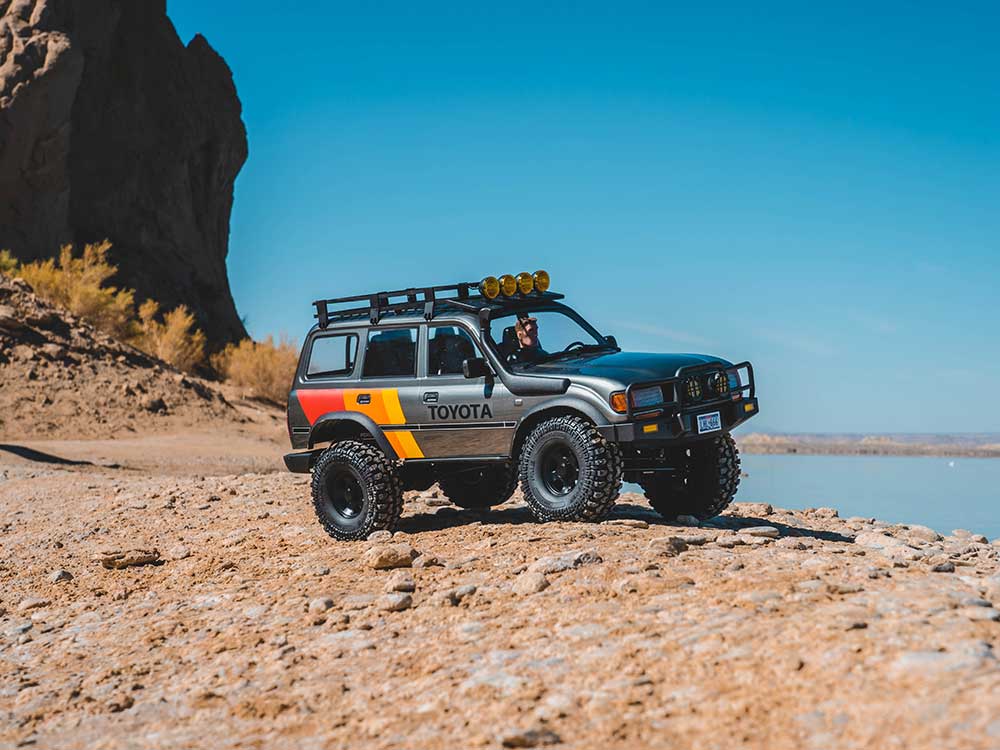 A miniature Toyota SUV model with off-road features, including large tires, a roof rack, and roof-mounted lights, placed on a sandy ground with a cliff and a lake in the background.