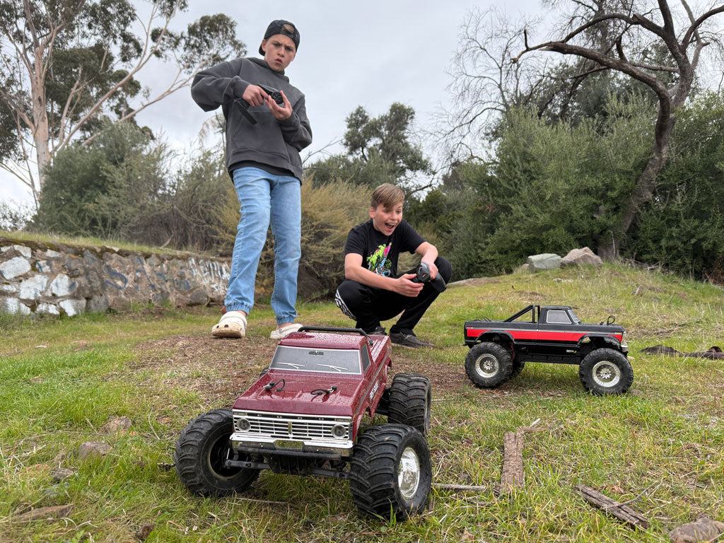 Dos chicos operan entusiastamente sus camiones RC Fmshobby en un campo de hierba, mostrando un rendimiento todoterreno detallado