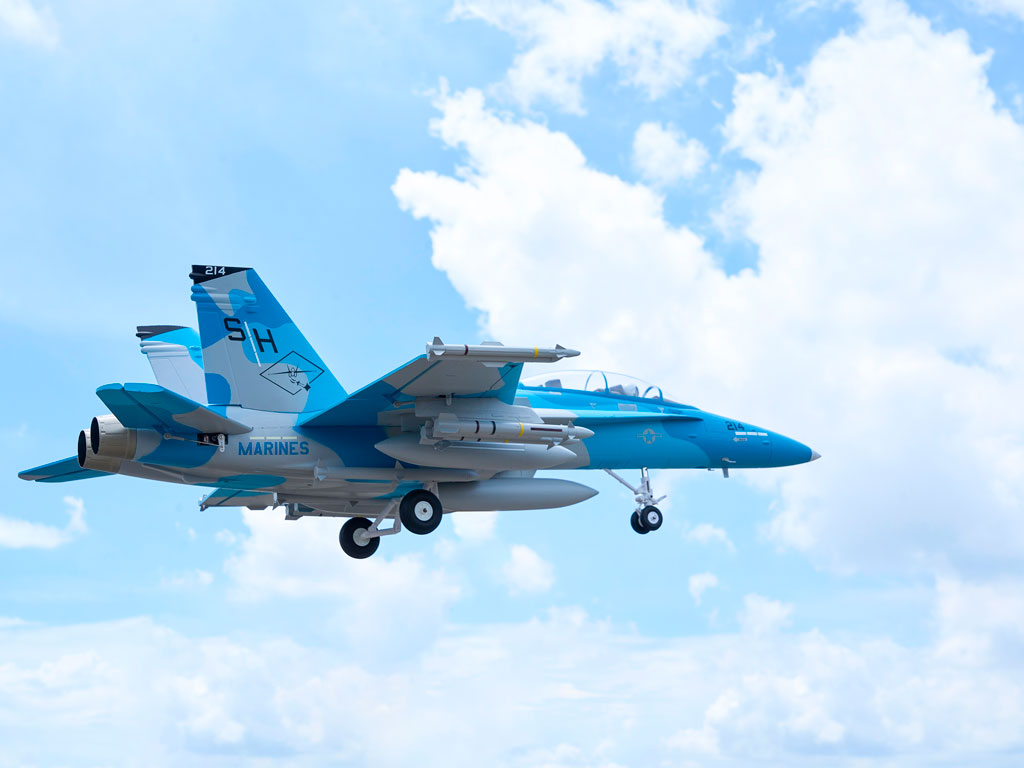 A blue and white RC jet airplane with "MARINES" and "SH" markings, flying against a bright sky with scattered clouds.