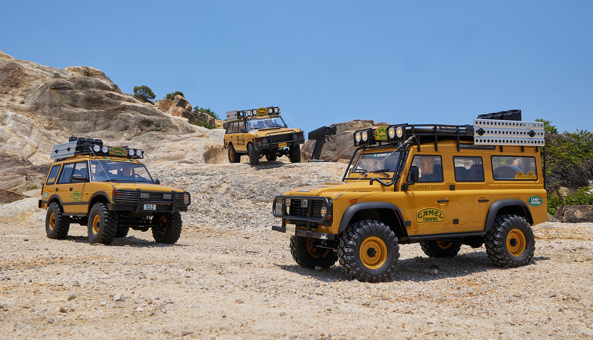 Three yellow Camel Trophy-themed miniature off-road vehicles with roof racks and expedition equipment, displayed on a rocky landscape under a clear blue sky.