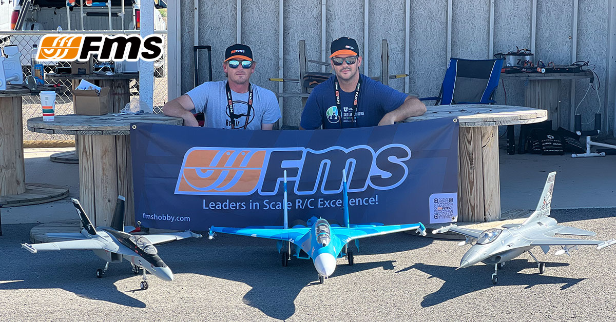 Two FMS representatives posing with three scale R/C fighter jet models in front of an FMS branded banner.