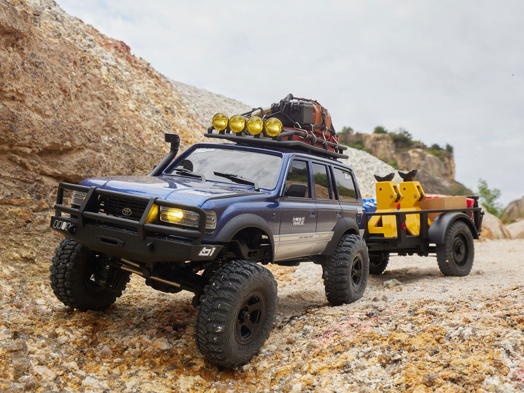 Un SUV RC Fmshobby azul transporta un remolque de equipo pesado a través de un camino montañoso accidentado