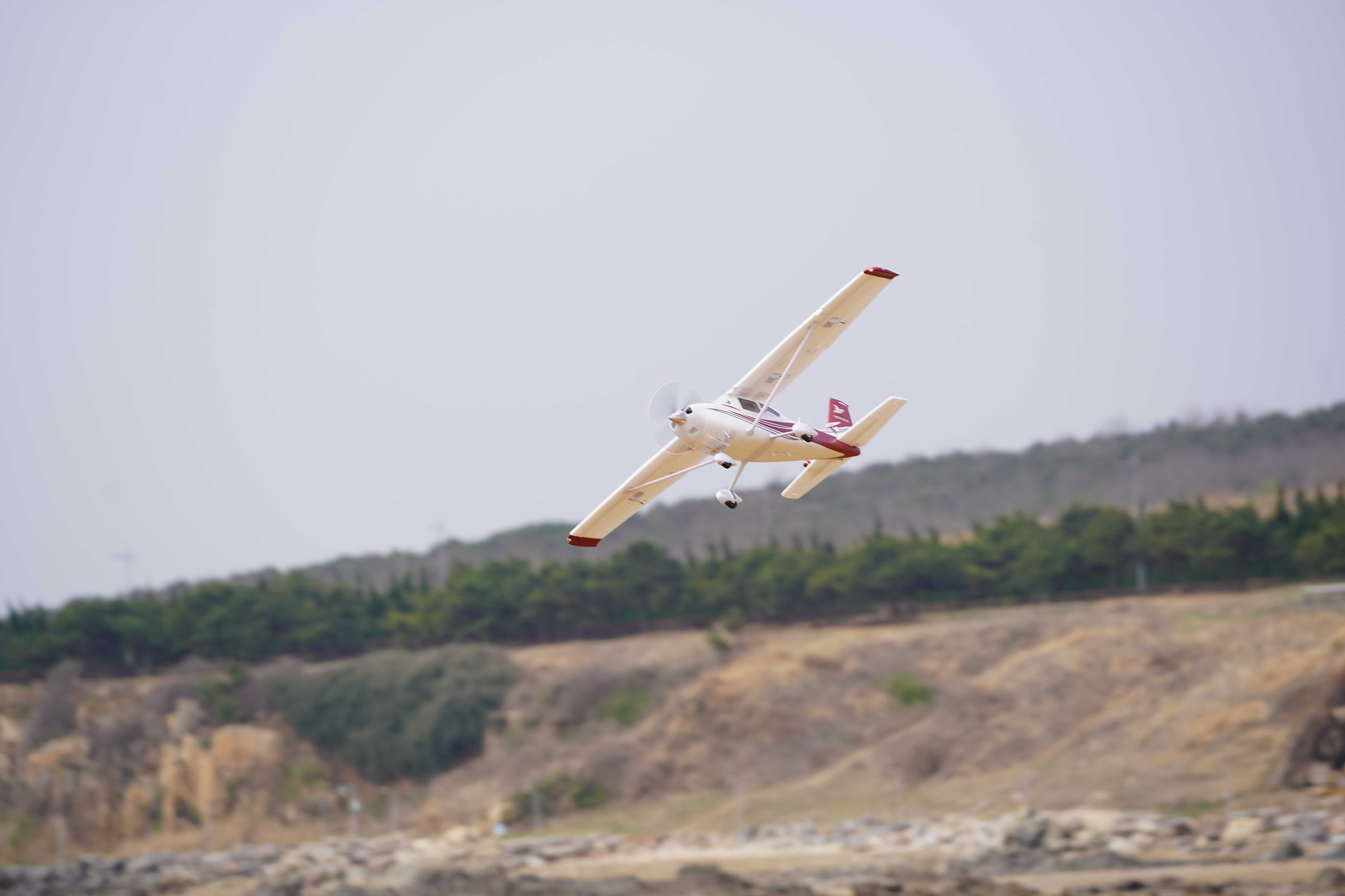 A white model airplane with red accents flying in the sky above a landscape.