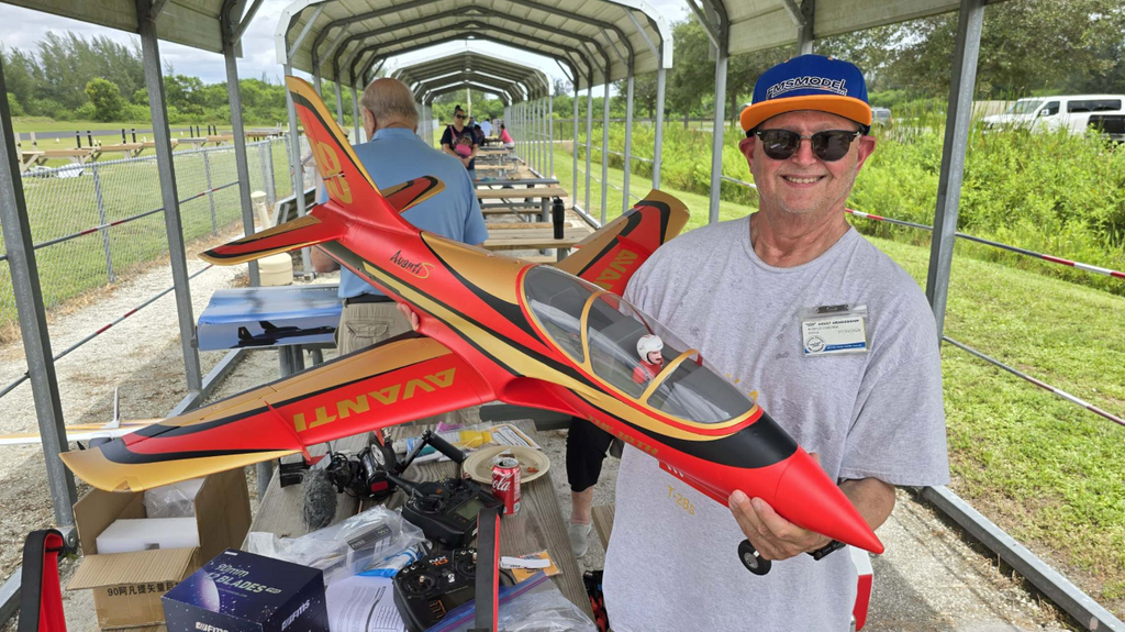 A man in a gray t-shirt 
and baseball cap smiles as he holds a remote-controlled model jet.