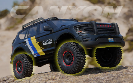 A rugged RC off-road vehicle labeled 'Canyon' with a bold black design, yellow and blue accents, and red-rimmed tires, parked on rocky terrain under a clear sky.