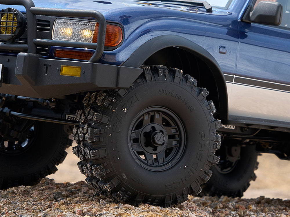A close-up view of an off-road tire on a miniature Toyota SUV model, showing detailed tread patterns and sidewall text, with a rocky ground surface in the background.
