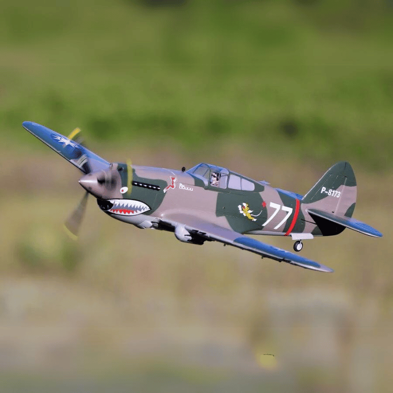 FMS 980mm P-40B Flying Tiger aircraft in flight, featuring shark mouth nose art and '77' marking.