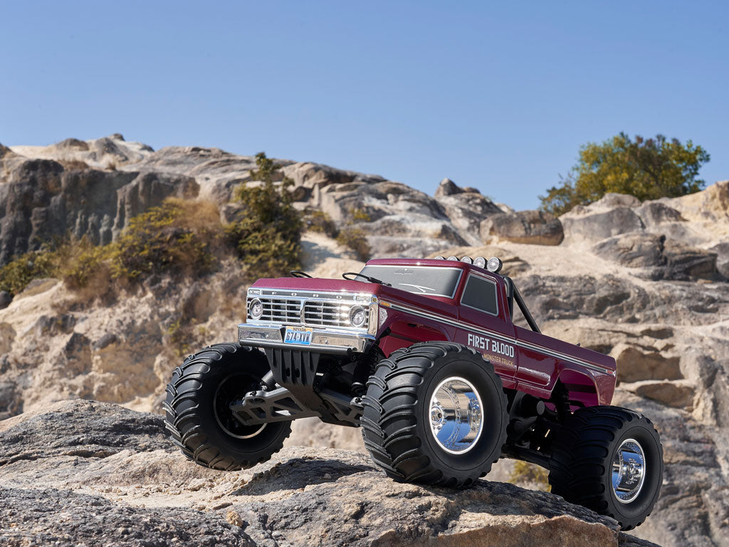 A maroon FMS RC Monster Truck with "First Blood" decals conquers a rocky ridge against a clear blue sky