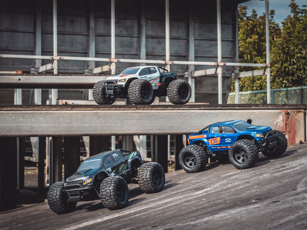 Three FMS RC Monster Truck models in white, blue, and black climb a steep concrete ramp at an industrial site