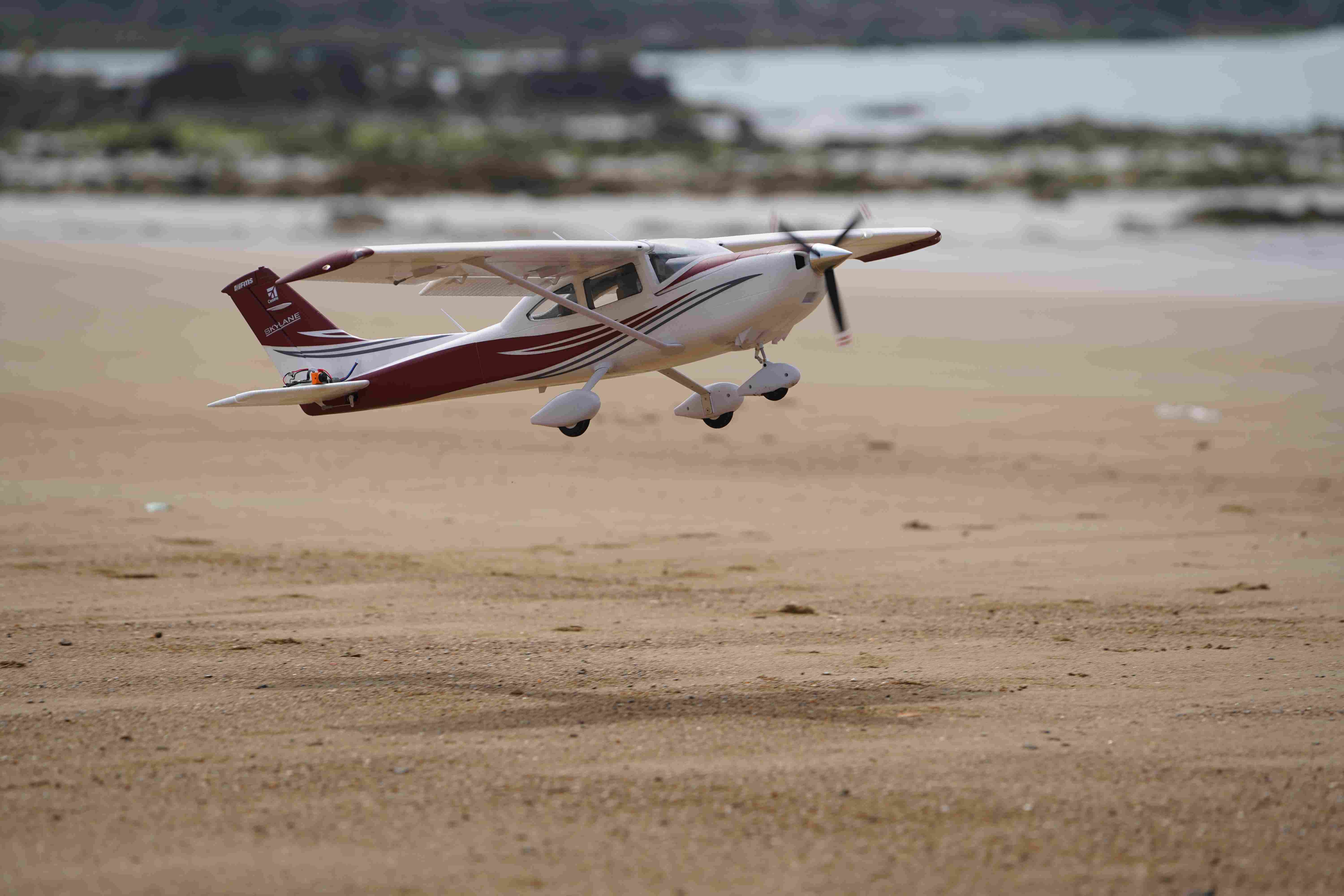 Fmshobby white maroon Skylane RC airplane model flying low over sandy beach with spinning propeller