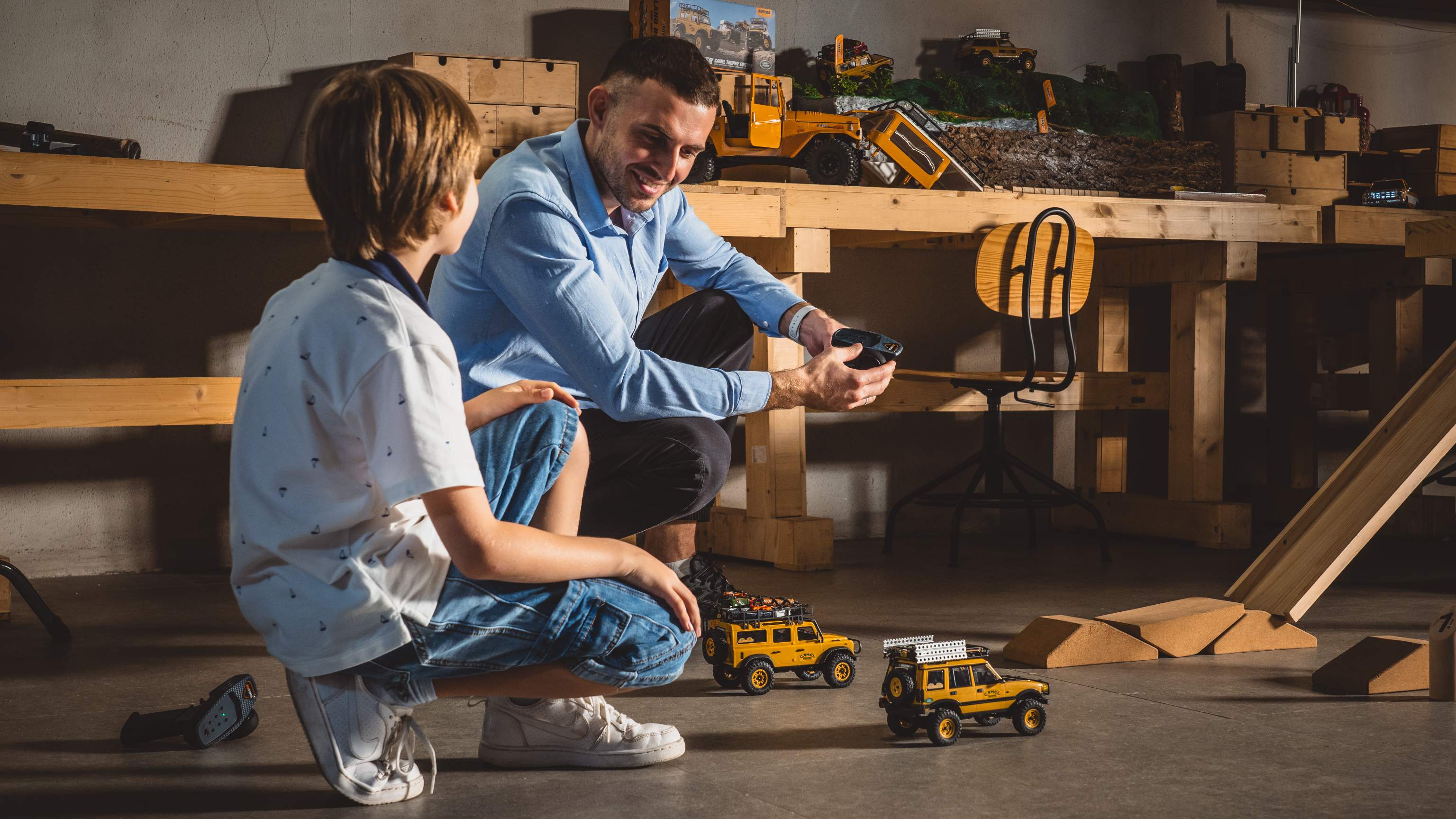 Adult and child driving two mini RC crawlers across a garage floor using a handheld transmitter
