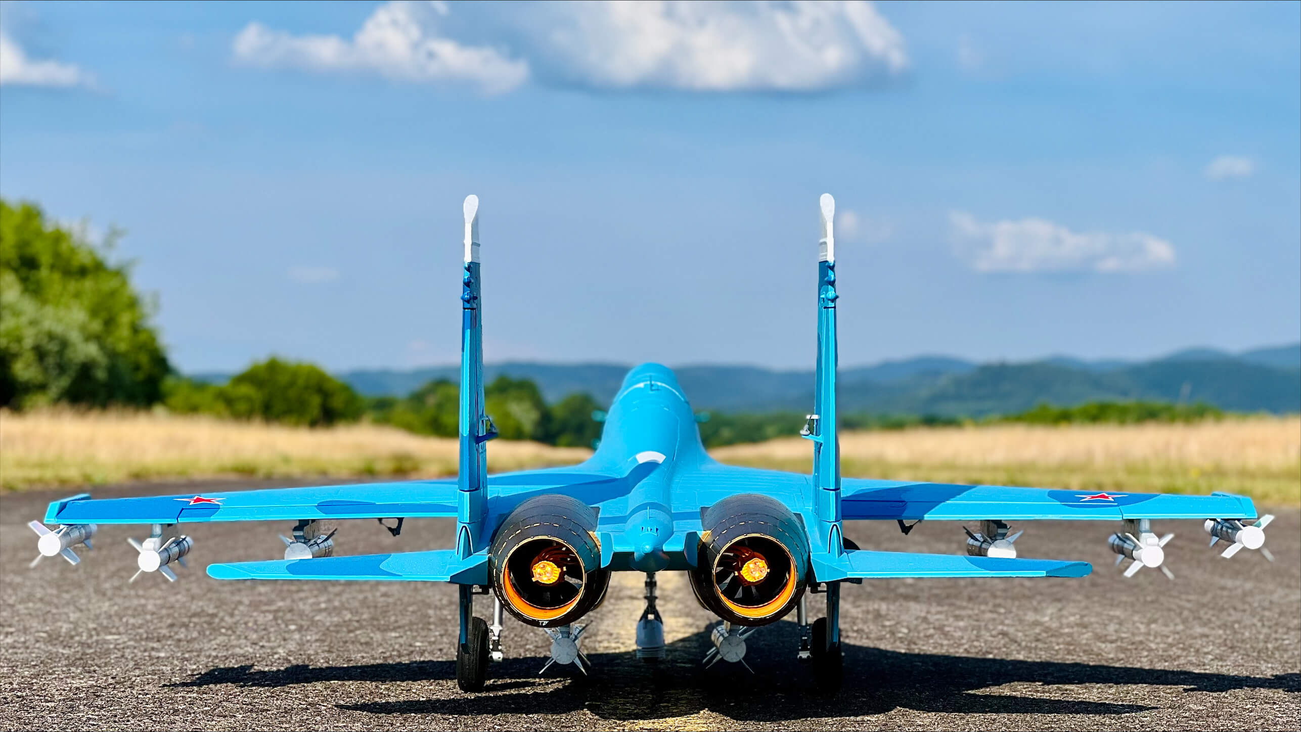 Rear view of a blue FMS scale fighter jet R/C model with detailed engine exhausts, sitting on a runway.