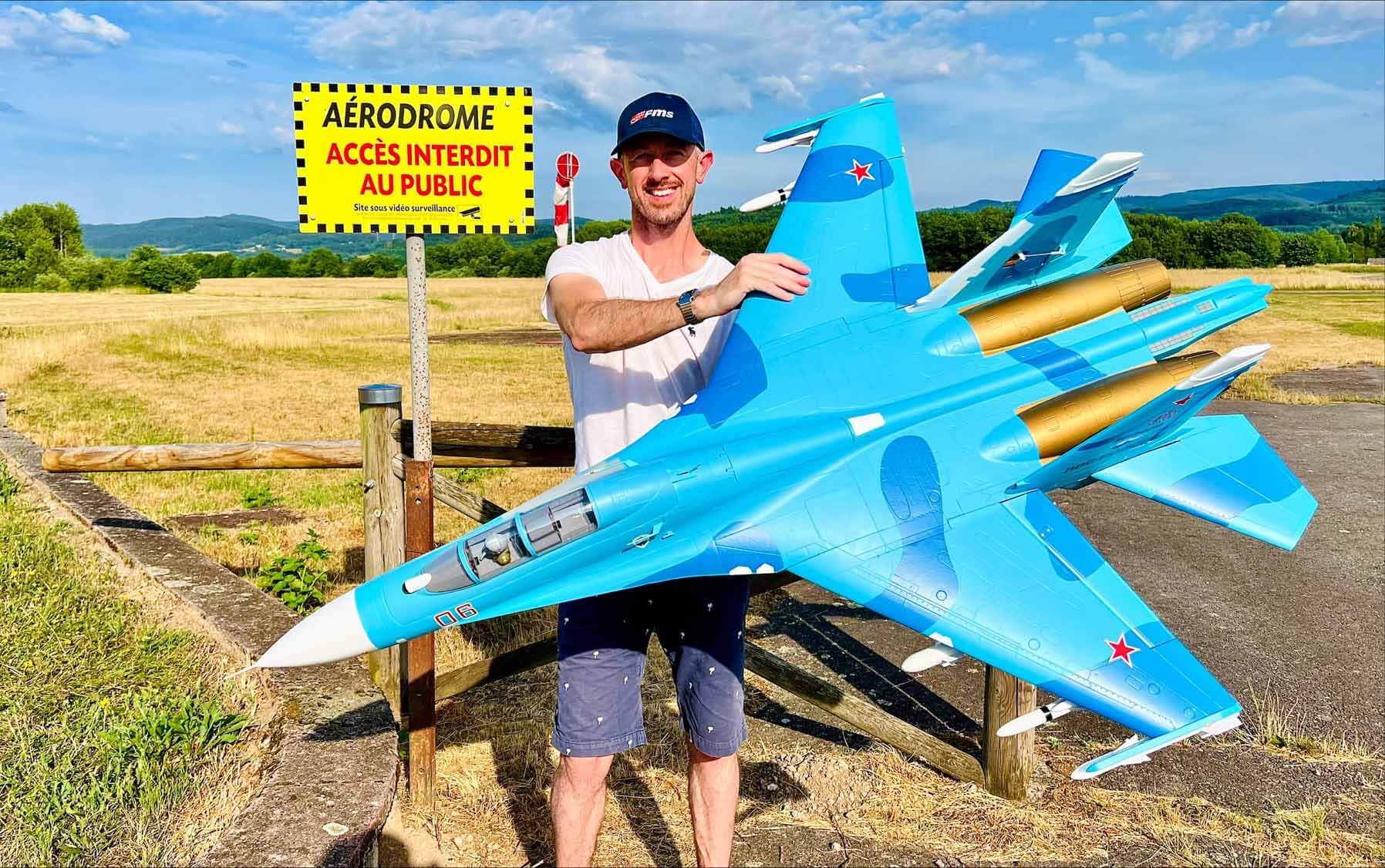 A man holding a large, blue camouflage FMS scale R/C fighter jet model at an airfield.