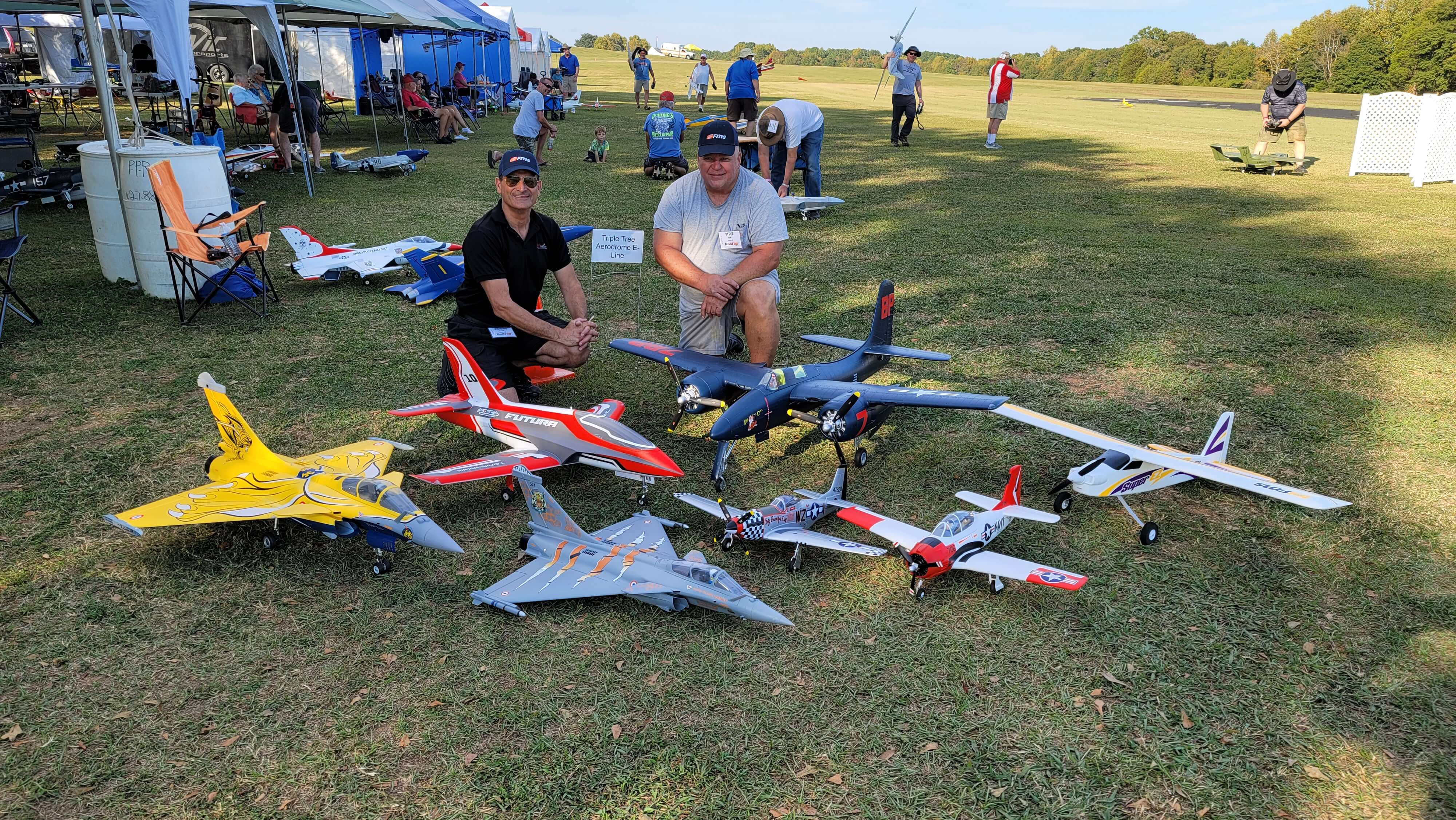 Two hobbyists posing with a collection of various FMS R/C airplanes at an outdoor airfield event.