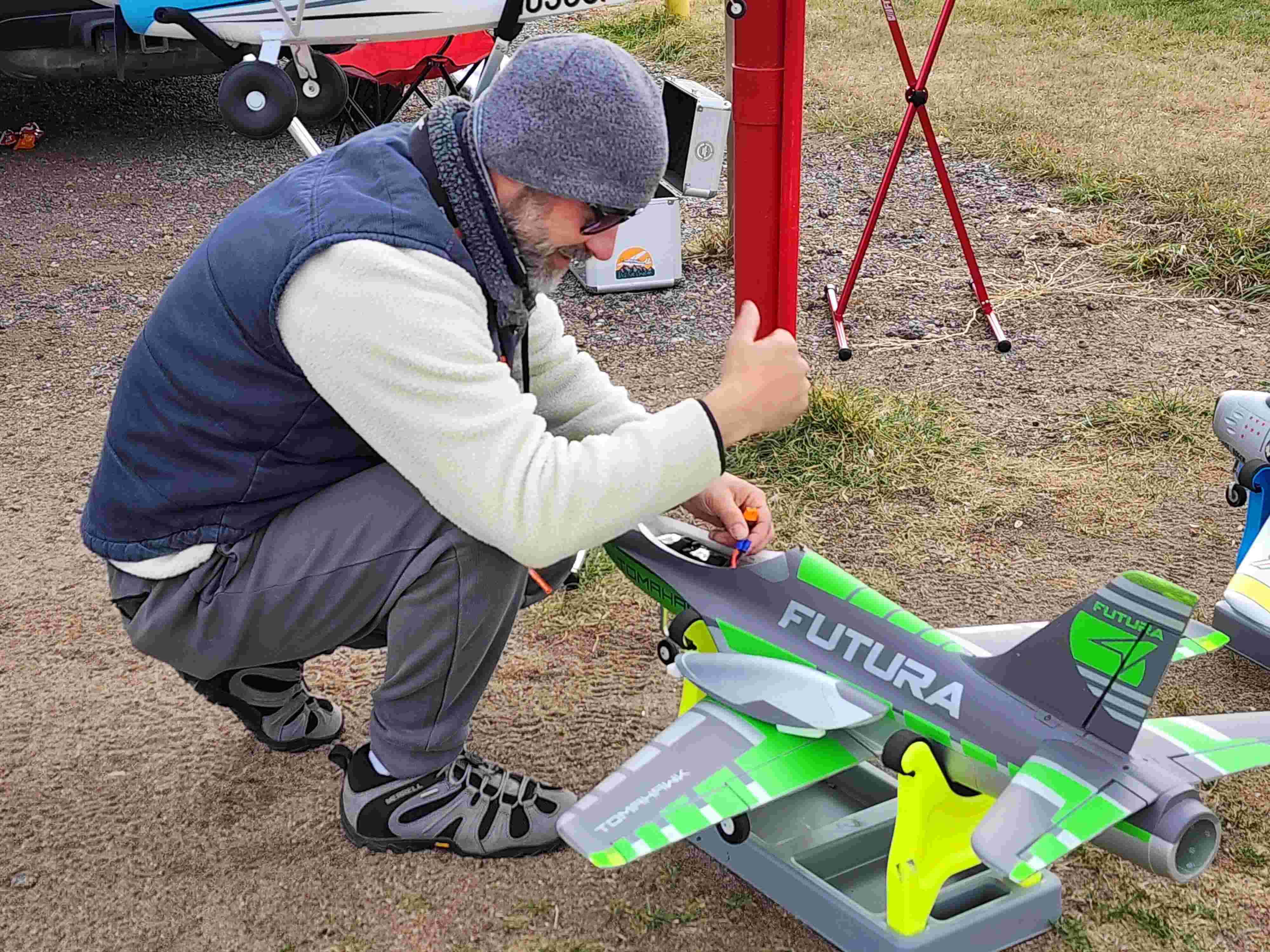 A pilot kneeling on the ground, preparing and checking the battery of his green and grey FMS Futura R/C jet model.