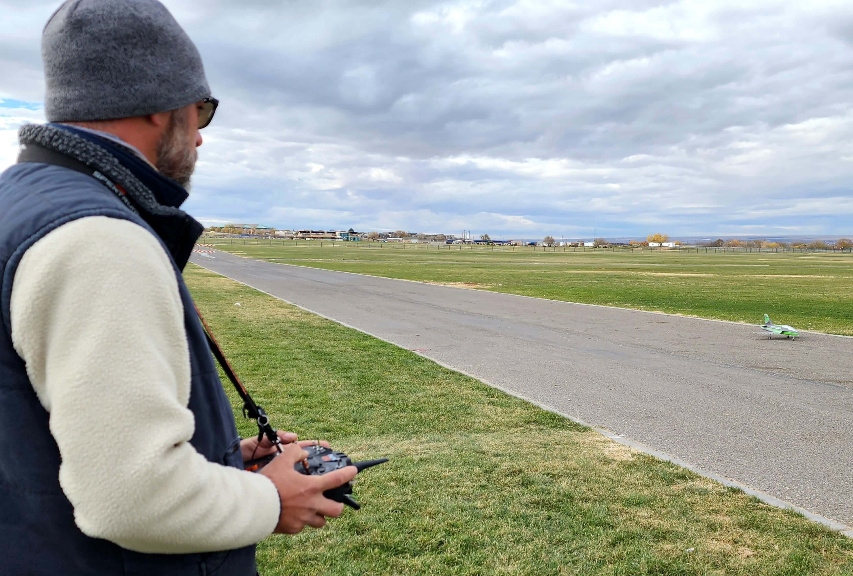 A pilot using a remote controller to fly an R/C airplane on a paved runway at an airfield.