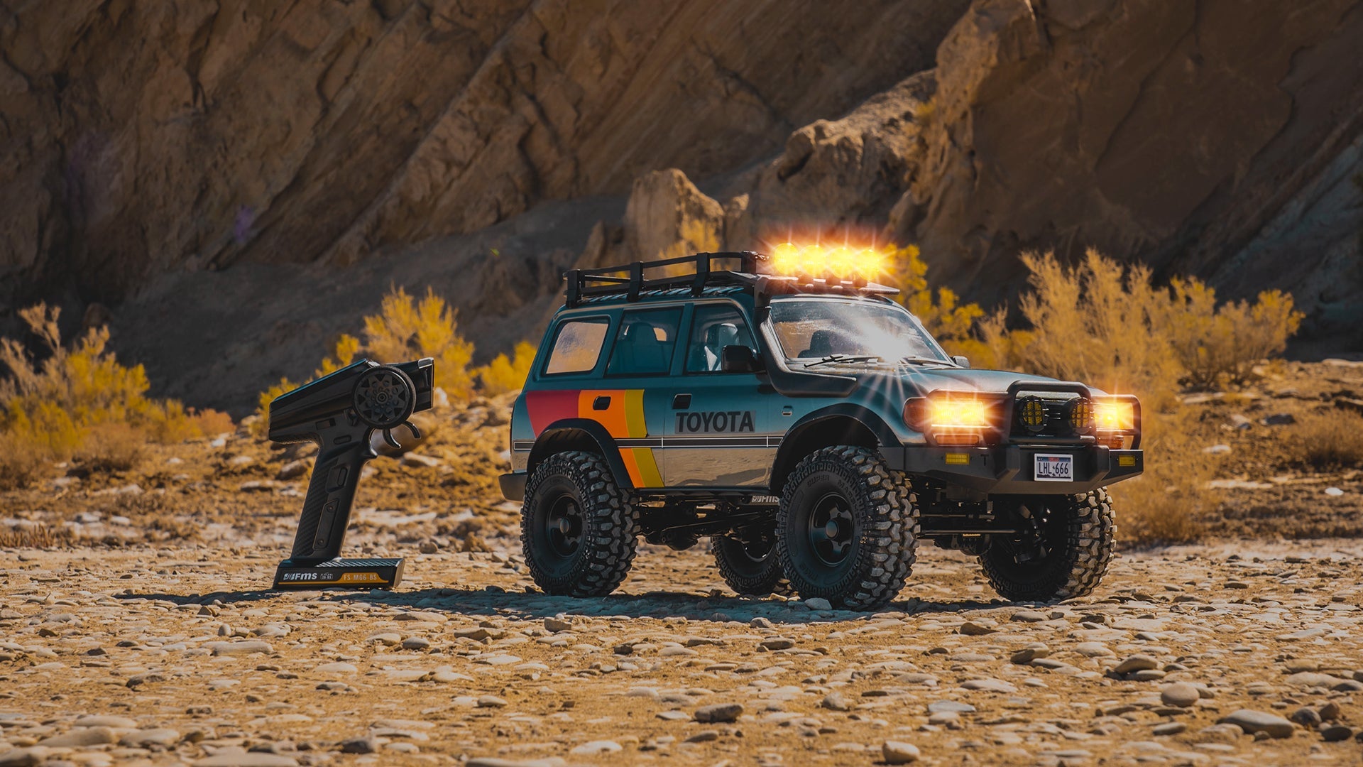 1/10-scale RC crawler with roof light bar lit, parked on desert gravel beside an FMS transmitter below a canyon wall