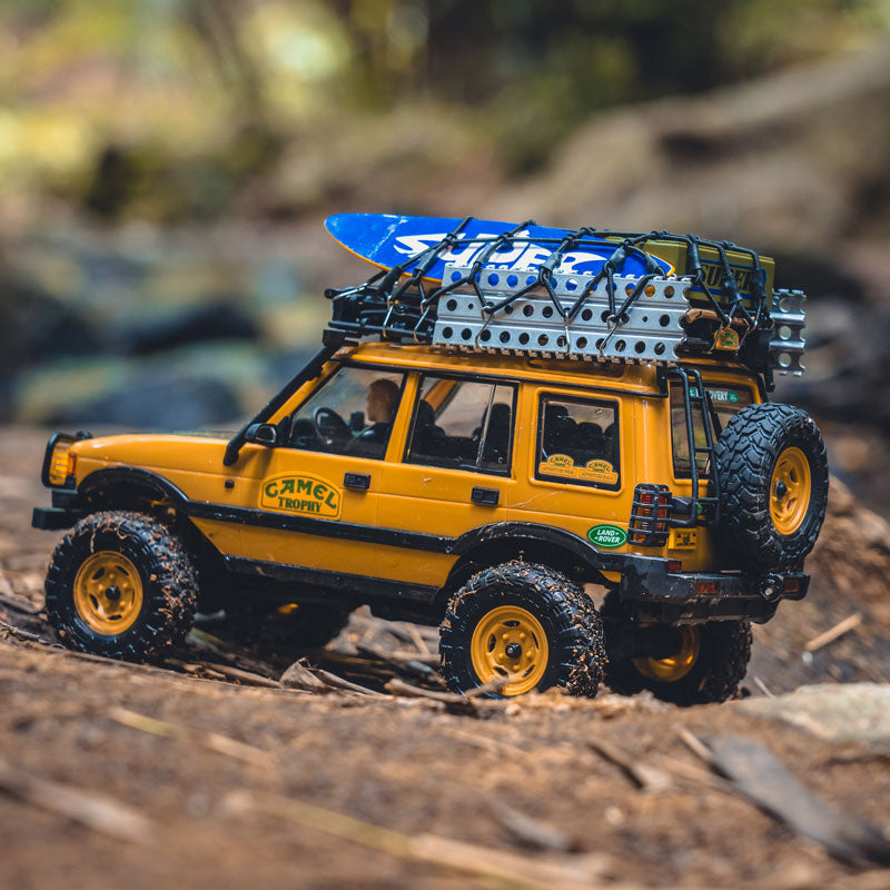 Yellow FMS 1:24 FCX24M Land Rover Discovery Camel Trophy RC crawler with surfboard on roof rack.