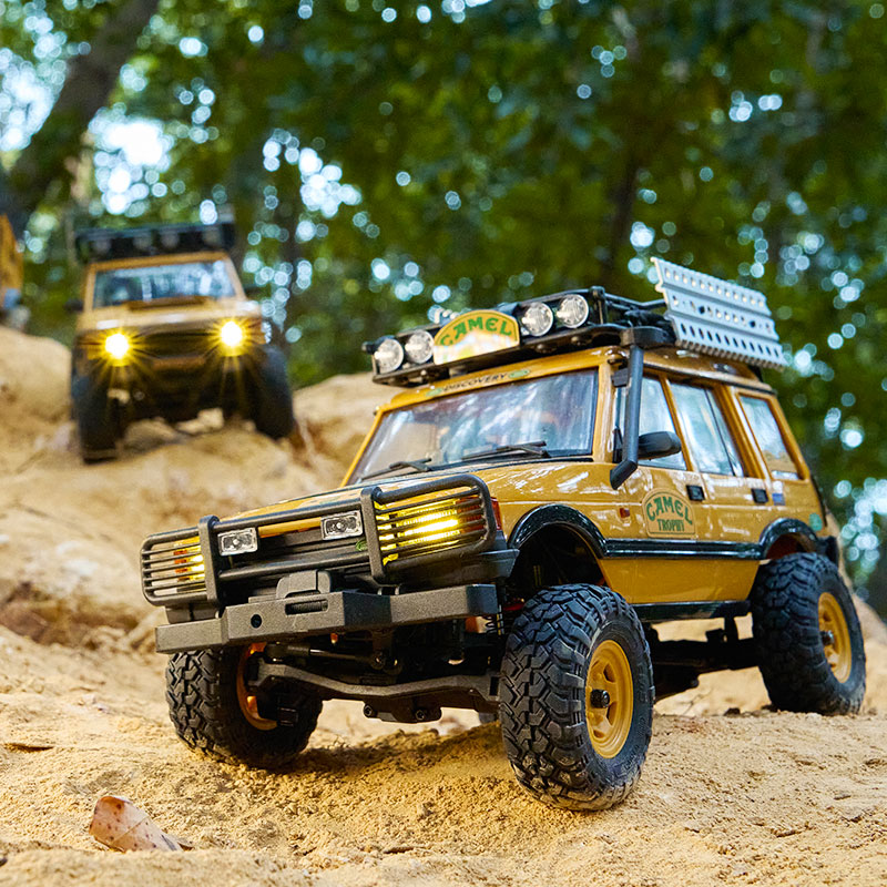 Two Camel Trophy-branded RC model cars with lights on, driving on a dirt trail.