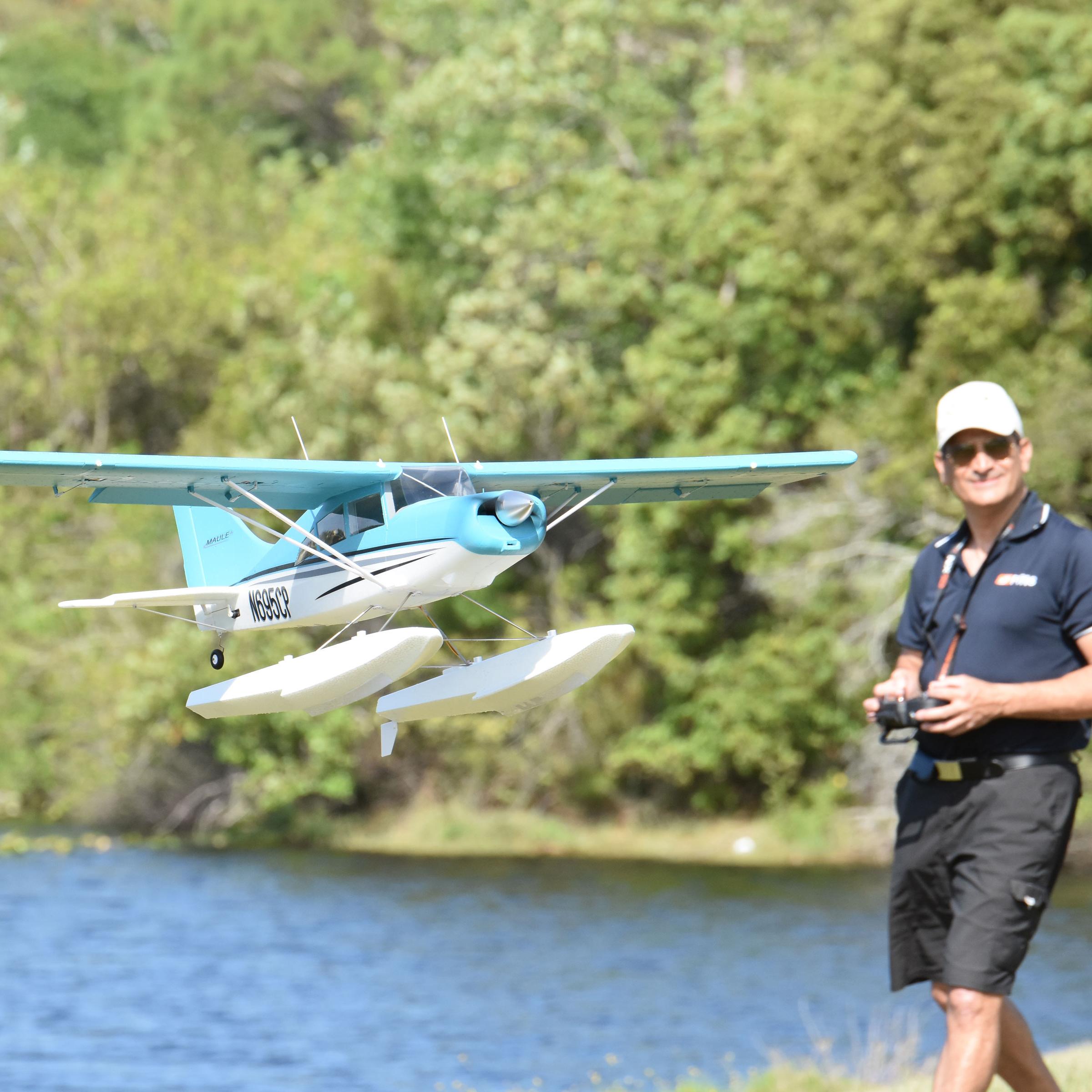 FMS 1400mm Kingfisher RC floatplane flying low over water with a pilot holding a transmitter