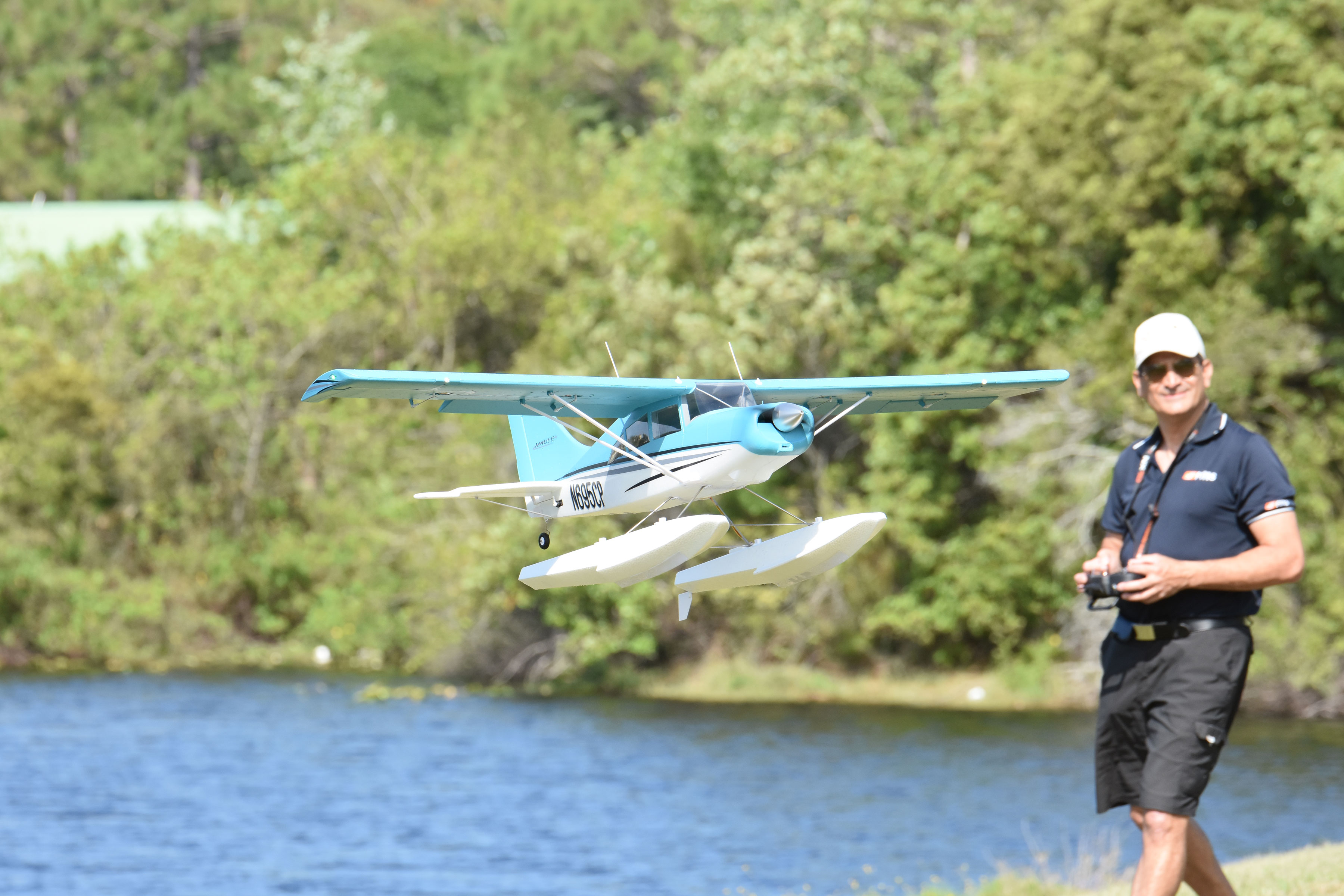 Fmshobby blue and white Maule RC seaplane on floats flying over water with pilot holding remote controller