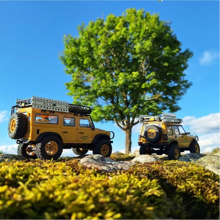 Two yellow FMS mini RC off-road trucks crawling over mossy rocks under a tree
