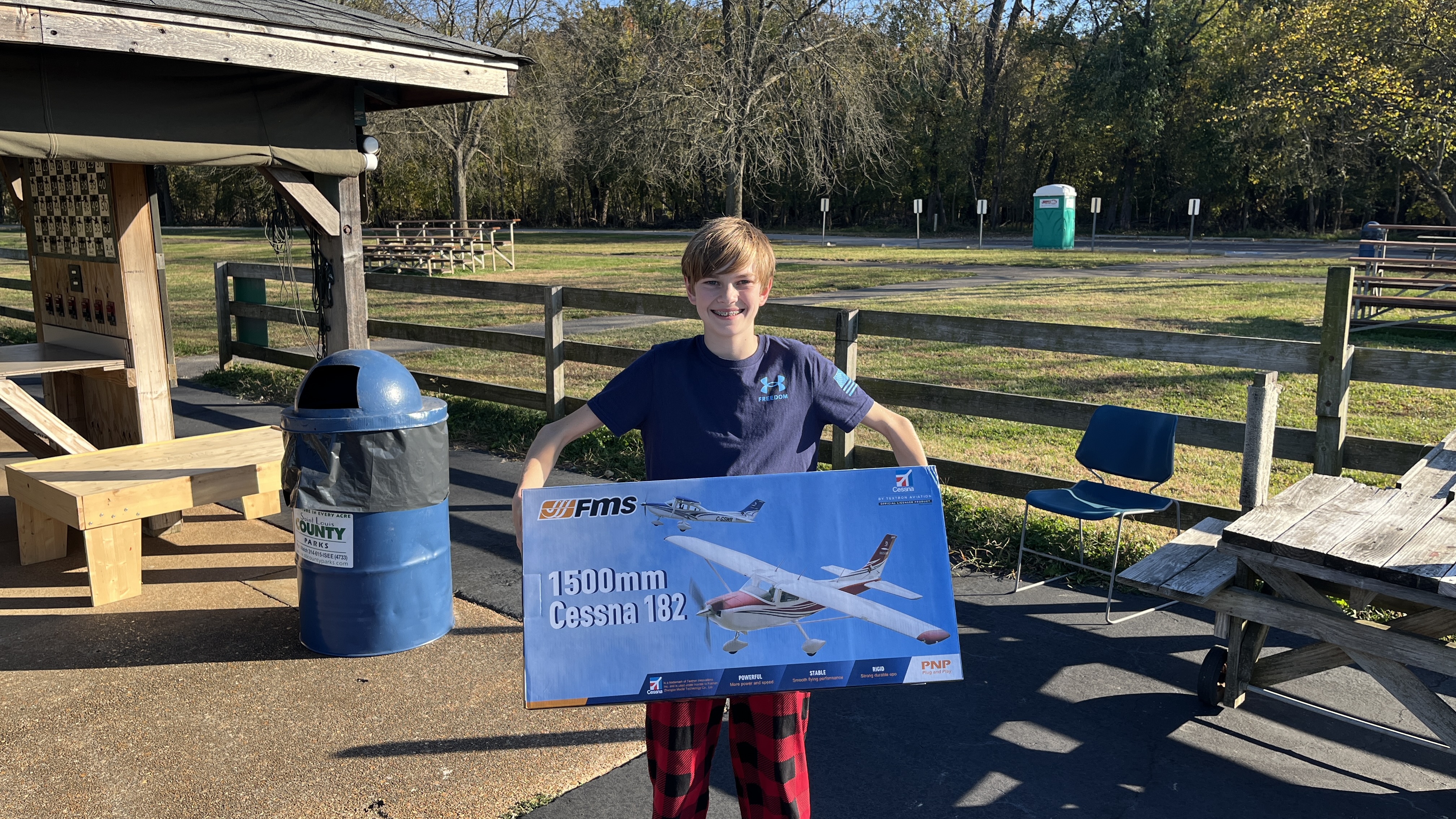 A young boy happily holding a boxed FMS 1500mm Cessna 182 R/C airplane kit at an outdoor park.