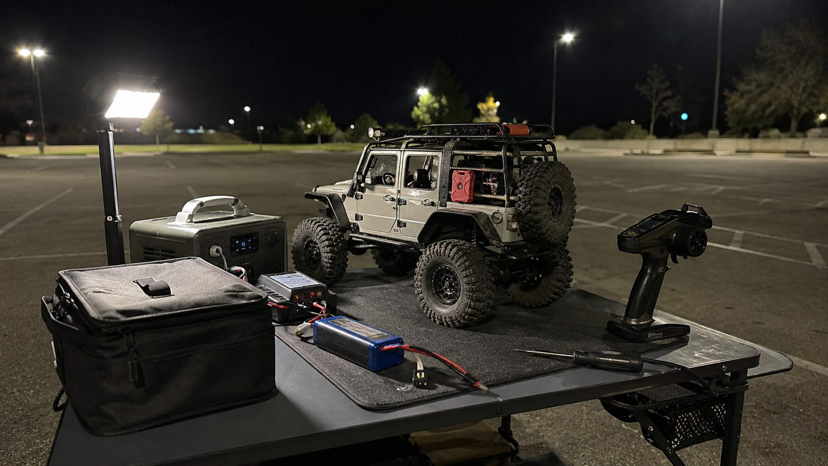 FMS RC rock crawler on a night pit table with transmitter, LiPo battery, charger, and portable work light