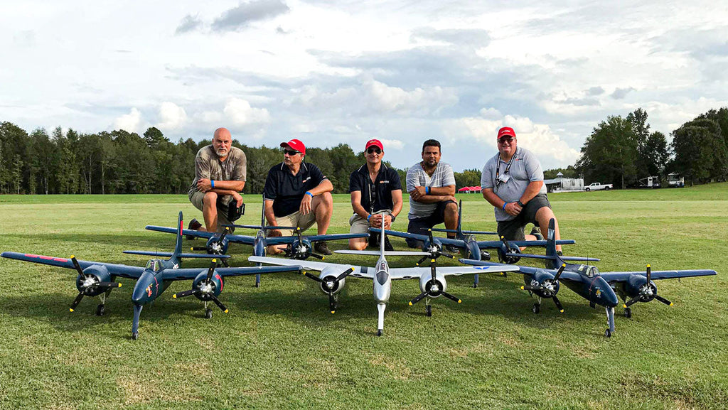Five men kneeling on grass behind five blue and white remote control model planes resembling WWII multi-engine fighters, with trees and cloudy sky in the background.